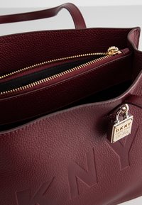 Burgundowa torba ręczna wykonana z teksturowanej skóry, z złotym zapięciem na zamek, czarną podszewką i metalowym akcentem logo DKNY.