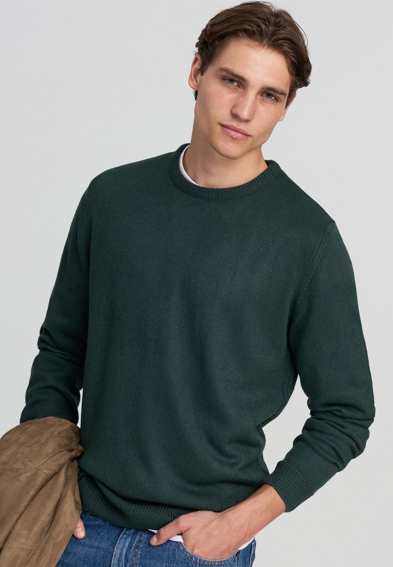 Maglione lavorato a maglia di colore verde scuro con scollatura rotonda e polsini a coste. Indossato con jeans di denim blu e una giacca in suede marrone appesa al braccio.
