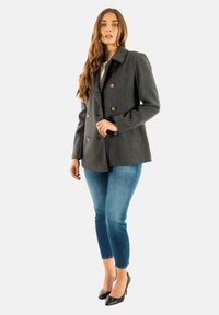 Cappotto grigio in misto lana doppiopetto con bottoni in ottone, grande colletto e tasche laterali, abbinato a jeans skinny blu e tacchi neri.