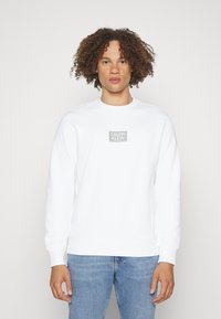 Felpa bianca con collo a costine, maniche lunghe e un piccolo logo grigio "Calvin Klein" sul petto, abbinata a jeans blu.