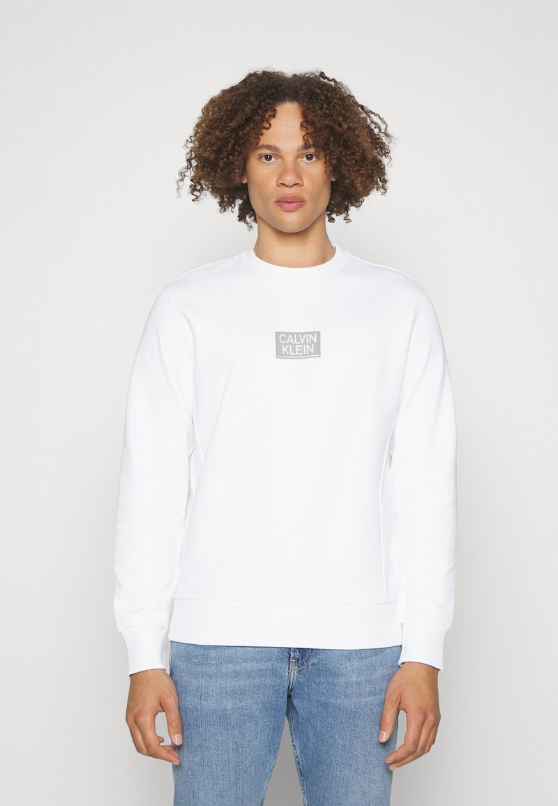 Felpa bianca con collo a costine, maniche lunghe e un piccolo logo grigio "Calvin Klein" sul petto, abbinata a jeans blu.