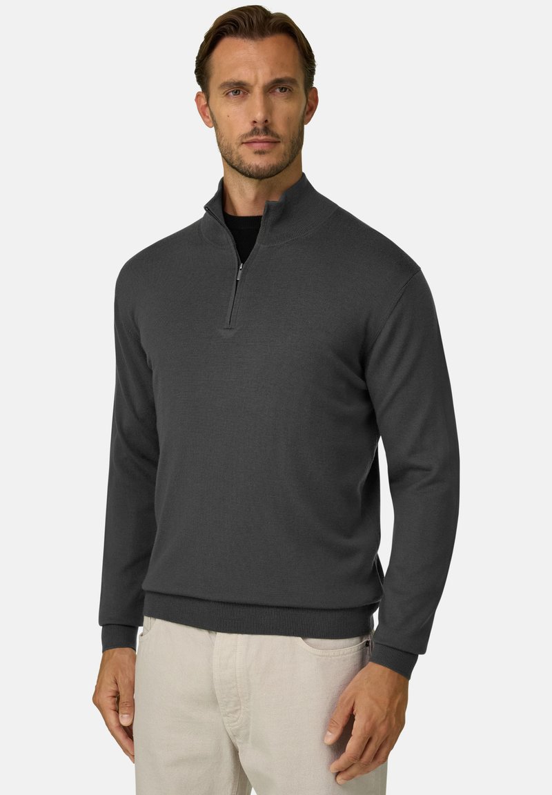 Homme portant un pull-over gris foncé à fermeture éclair quart de zip et un pantalon beige clair, debout devant un fond clair uni.