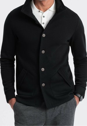 Homme portant une veste noire boutonnée sur une chemise blanche à col, un pantalon gris et une montre au poignet, les mains dans les poches.