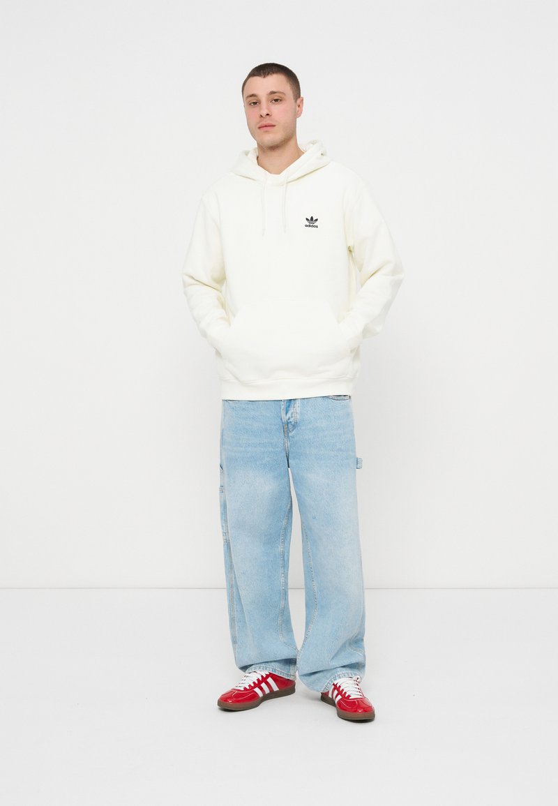 Młody mężczyzna stojący z rękami w kieszeniach, ubrany w białą bluzę z kapturem Adidas, jasnoniebieskie spodnie jeansowe o szerokich nogawkach oraz czerwone buty sportowe Adidas.