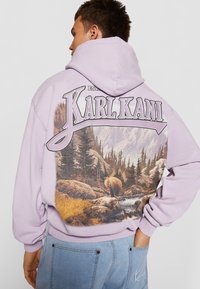 Hoodie lavanda con una grafica di paesaggio forestale sul retro, con il testo "KARL KANI" sopra. Materiale in cotone con polsini e orlo a costine.