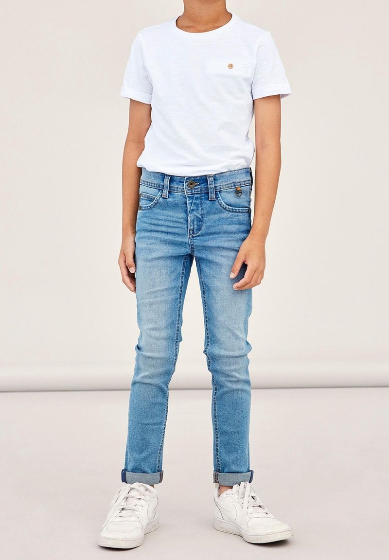 Enfant portant un t-shirt blanc uni à manches courtes, un jean bleu clair retroussé et des baskets blanches, debout devant un fond neutre.
