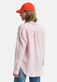 Camicia a maniche lunghe a righe rosa e bianche, con vestibilità comoda, polsini con bottone e una piega sul retro. Abbinata a un cappellino rosso vivo.