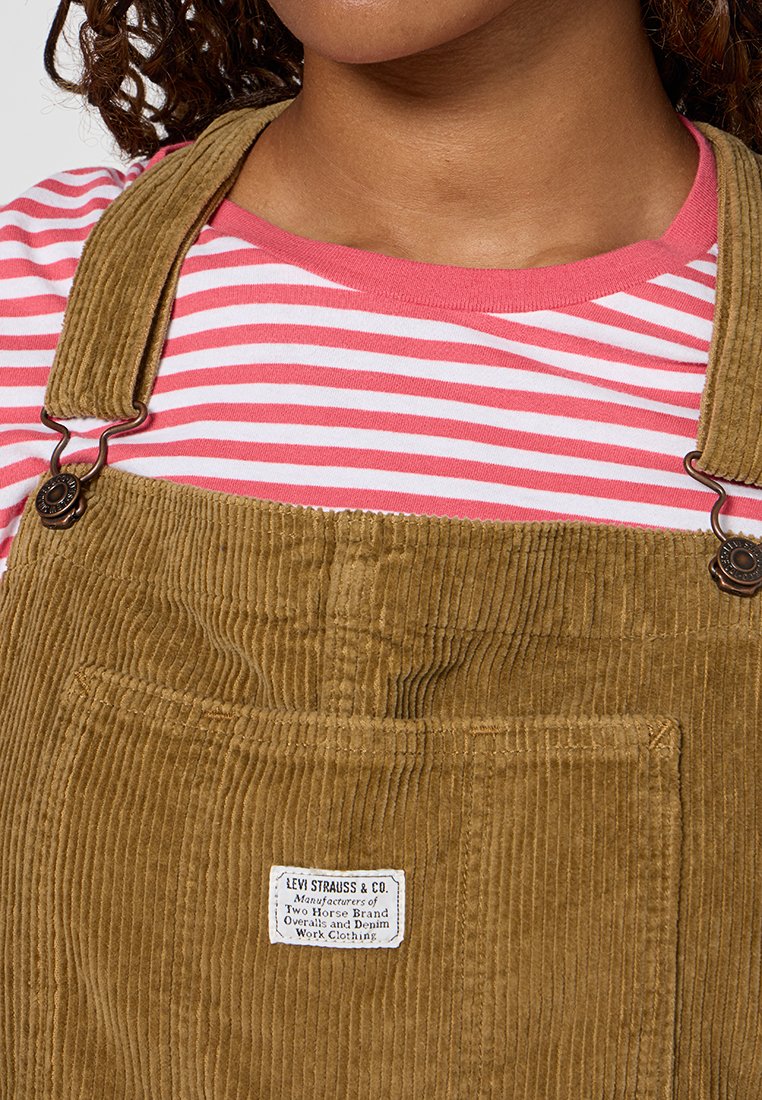 Salopette en velours côtelé de couleur brun clair, dotée de clips métalliques réglables et d'une poche frontale. Portée par-dessus une chemise à rayures roses et blanches.