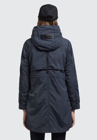 Garšs, tumši zils parka mētelis ar kapuci un rāvējslēdzēju, ar strukturētu aizmugures paneli un logotipa akcentu uz kapuces. Gluda auduma.