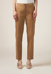 Pantaloni beige su misura con una vestibilità slim, caratterizzati da chiusura con bottone frontale, tasche laterali e una trama liscia. Indossati con scarpe di colore chiaro.