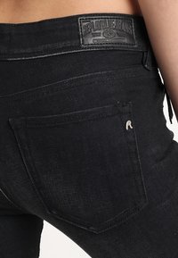 Jeans de mezclilla negra que presentan un primer plano del bolsillo trasero y la etiqueta de cuero en la cintura con texto grabado. La tela tiene una textura suave.