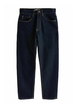 Femme portant un cardigan boutonné vert foncé, un jean bleu foncé et des chaussures à talons bordeaux, debout sur un fond marron.