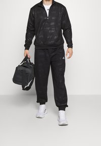 Černá tepláková souprava s bundou na zip a jemným potiskem loga, doplněná kalhotami. K tomu šedá sportovní taška a bílé boty.