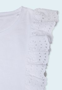 Vit t-shirt med rund halsringning som har en volangspets detalj på axeln, texturerad med utskurna mönster och ett slätt tyg.