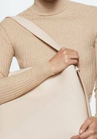 Sac à épaule en cuir beige avec une texture lisse et une large bandoulière ajustable, tenu par une personne portant un pull beige à côtes.