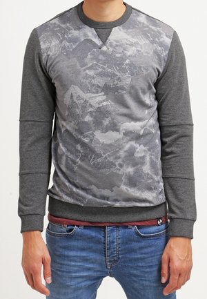 Mand iført en grå sweatshirt med et bjergmotiv foran, kombineret med blå jeans og en rød skjorte under.