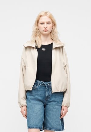 Ung kvinne med blondt hår iført en beige jakke med glidelås over en svart topp og blå jeansshorts, stående foran en ensfarget bakgrunn.