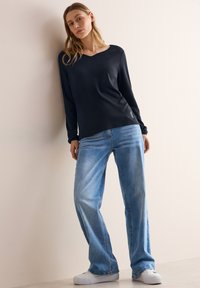 Top navy a maniche lunghe con scollo a V abbinato a jeans dritti azzurri e sneakers bianche, con un discreto dettaglio del logo sul top.
