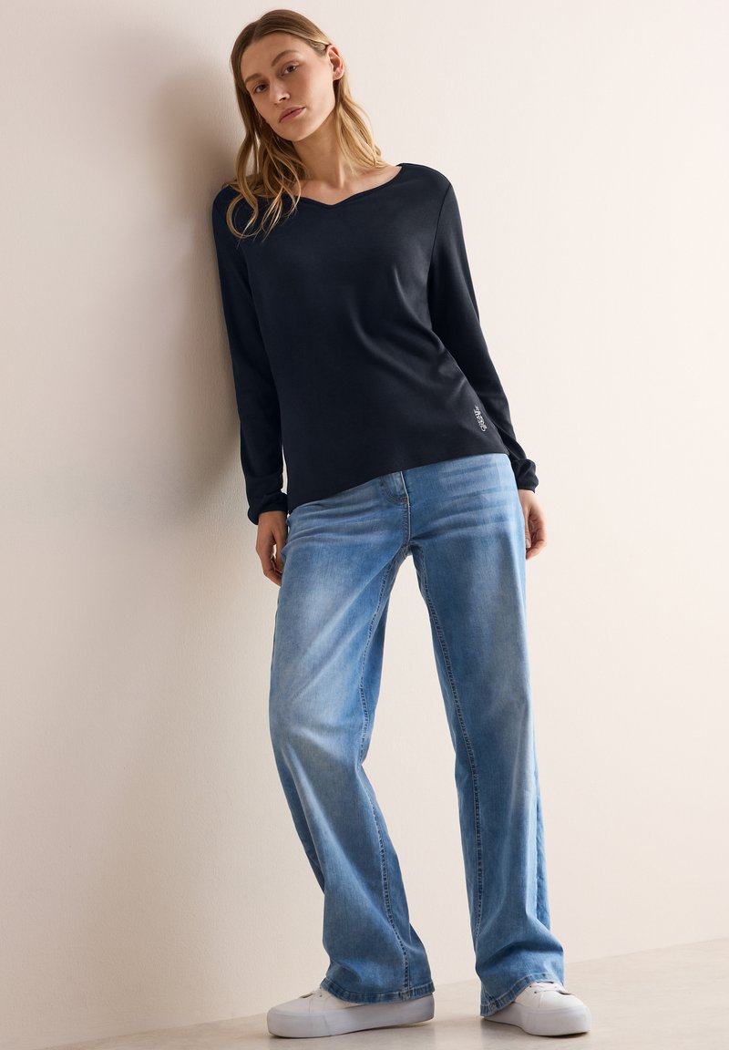 Top navy a maniche lunghe con scollo a V abbinato a jeans dritti azzurri e sneakers bianche, con un discreto dettaglio del logo sul top.