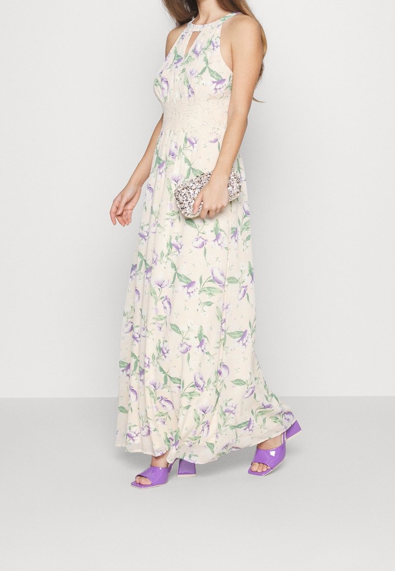 Femme portant une robe longue crème sans manches avec un imprimé floral violet, tenant une pochette scintillante et portant des sandales à talons violettes.