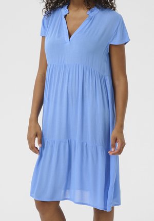 Mujer con vestido azul claro de manga corta, largo hasta la rodilla, con cuello en V con volantes y cintura y paneles de falda fruncidos.