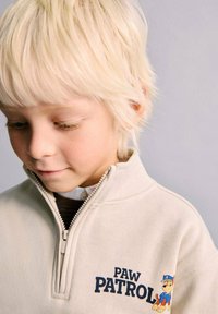 Bež pulover s kapuco in zadrgo iz flisa z napisom "PAW PATROL" in vezenim likom. Mehka tekstura, sproščen kroj, kontrastni temno modri poudarki.
