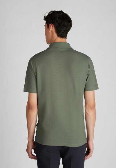 Homme portant un polo à manches courtes olive et un pantalon sombre, se tenant de dos contre un fond clair.