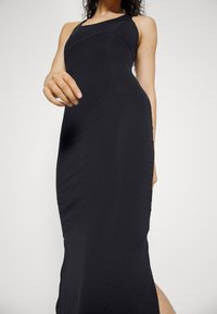 Vestido preto acanalado com uma silhueta ajustada, alças finas e uma fenda lateral, apresentando um tecido texturizado e um design minimalista.