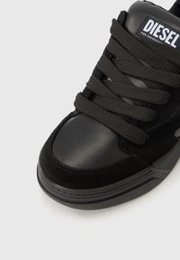 Μαύρο αθλητικό παπούτσι που συνδυάζει λείο δέρμα και υφή σουέτ, στρογγυλή μύτη, χοντρές κορδόνια και ετικέτα με το λογότυπο στη γλώσσα.