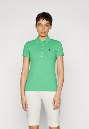 Green cotton polo shirt with short sleeves, a collar, and five buttons. Features a dark blue logo on the left chest. Worn with white shorts.