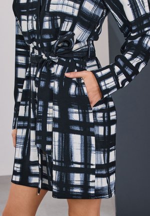 Zwart-wit geruite jurk met ceintuur en zakken, met een hand die rust in de rechterzak.