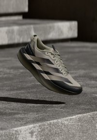Beiger Sportschuh mit schwarzen Akzenten, strukturierter Sohle und stromlinienförmigem Design. Verfügt über Schnürsenkel und ein Logo an der Seite, schwebt über einer Betonoberfläche.