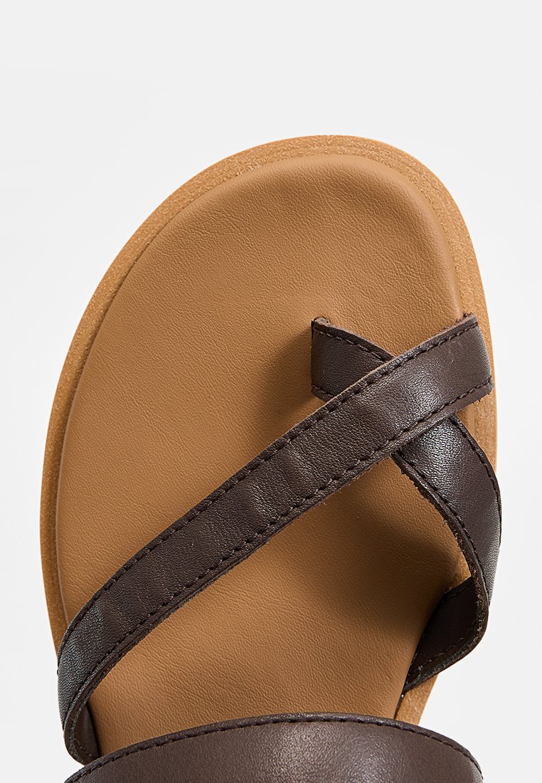 Sandalia de cuero marrón con suela plana, que presenta tiras cruzadas y una plantilla lisa. Los acentos texturizados en las tiras añaden detalle.