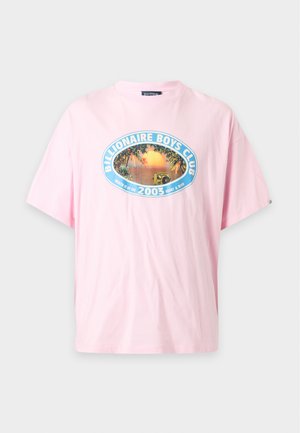Homme debout portant un t-shirt graphique rose oversize, un short en jean beige effiloché, des chaussettes blanches et des mocassins marron foncé sur fond blanc.