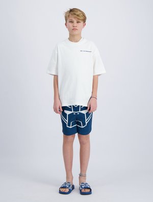 Blond jongetje met een oversized wit T-shirt, donkerblauwe short met geometrische print en bijpassende blauwe slippers, staand tegen een effen witte achtergrond.