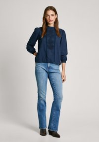 Blusa a maniche lunghe blu navy con maniche a sbuffo, bordo in pizzo e dettagli ricamati, abbinata a jeans blu chiaro e stivali neri con tacco.