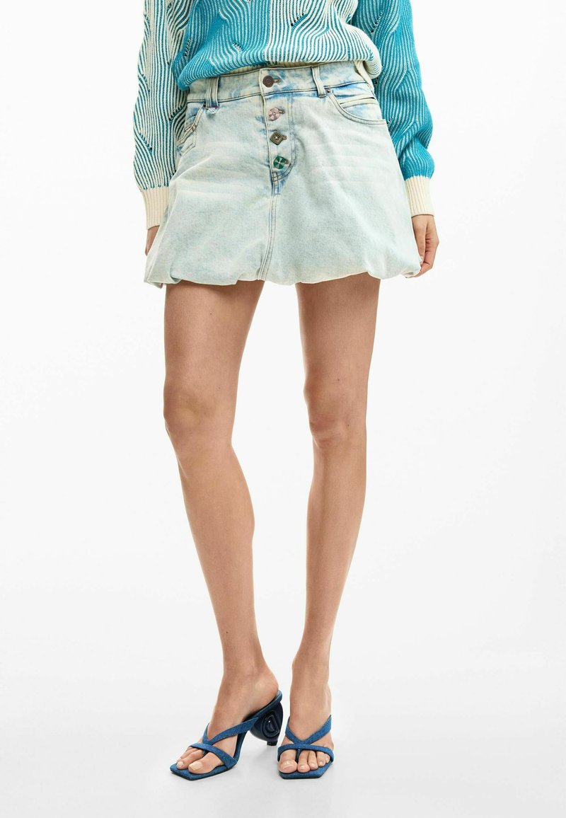 Persoană îmbrăcată într-un fustă mini din denim bleu deschis cu nasturi, sandale cu toc albastre cu barete și un pulover în dungi albastre și albe.