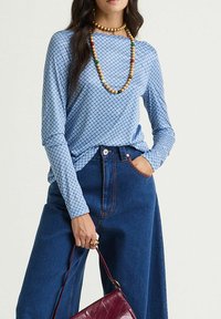 Haut bleu à manches longues avec un motif géométrique; associé à un pantalon en denim bleu taille haute; accessoirisé avec des colliers de perles colorées et un sac bordeaux.