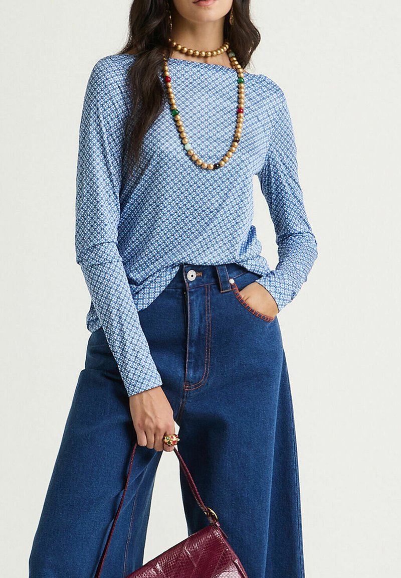 Haut bleu à manches longues avec un motif géométrique; associé à un pantalon en denim bleu taille haute; accessoirisé avec des colliers de perles colorées et un sac bordeaux.