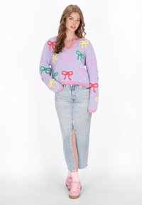 Lila gestrickter Pullover mit mehrfarbigen Schleifenmustern, kombiniert mit einem hellblauen Jeansrock mit einem Schlitz vorne und rosa Sneakers.