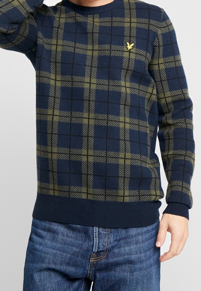 Pull en tricot à carreaux navy et olive avec des manches longues, un col rond, un ourlet côtelé et un petit logo jaune sur la poitrine.