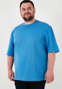 Modrá bavlnená tričko s okrúhlym výstrihom a krátkymi rukávmi. Vyznačuje sa voľným strihom a hladkou textúrou. Nosené s tmavými nohavicami.