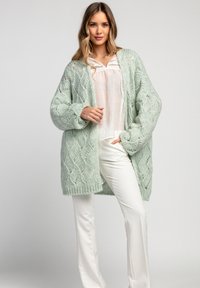 Miętowo-zielony sweter z dzianiny o teksturowanym, otwartym wzorze, noszony na białej bluzce i zestawiony z białymi spodniami. Długi, oversize'owy krój.