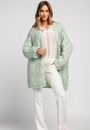 Miętowo-zielony sweter z dzianiny o teksturowanym, otwartym wzorze, noszony na białej bluzce i zestawiony z białymi spodniami. Długi, oversize'owy krój.