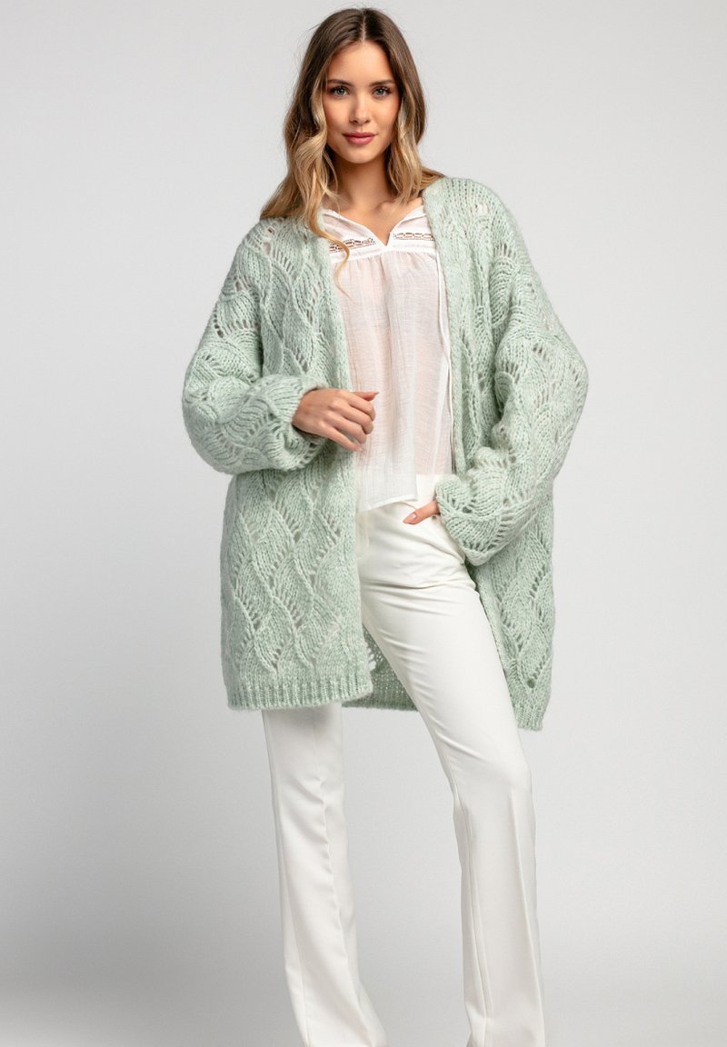 Miętowo-zielony sweter z dzianiny o teksturowanym, otwartym wzorze, noszony na białej bluzce i zestawiony z białymi spodniami. Długi, oversize'owy krój.