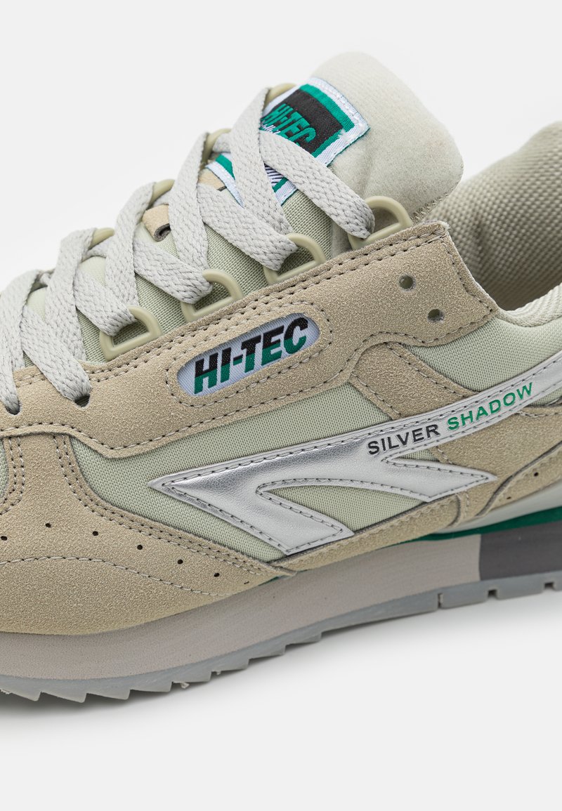 Beżowe zamszowe i materiałowe sneakersy z srebrnymi akcentami, z płaskimi sznurowadłami, perforacjami dla lepszej wentylacji oraz zielonym akcentem na śródpodeszwie.