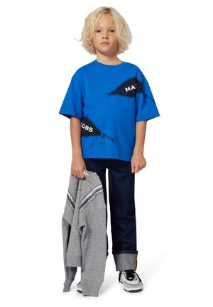 T-shirt bleu à manches courtes avec motif de fermeture éclair et texte noir ; associé à un jean en denim sombre et un sweat-shirt gris. Baskets blanches et noires.