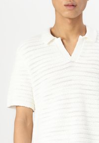Camisa polo blanca de manga corta con un patrón de punto texturizado, que presenta un cuello en V y un diseño de collar suave y casual.