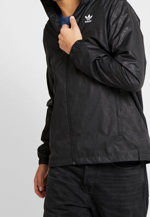 Mężczyzna w czarnej kurtce na zamek Adidas z subtelnym wzorem i białym logo, trzymający kołnierz przy szyi, w połączeniu z czarnymi dżinsami.