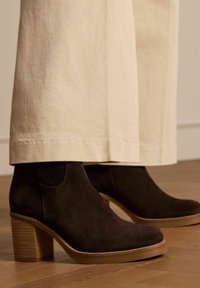 Donkerbruin suède enkellaarsjes met een chunky houten hak en een gladde rubberen zool. Ze hebben subtiele stiksels langs de bovenrand.